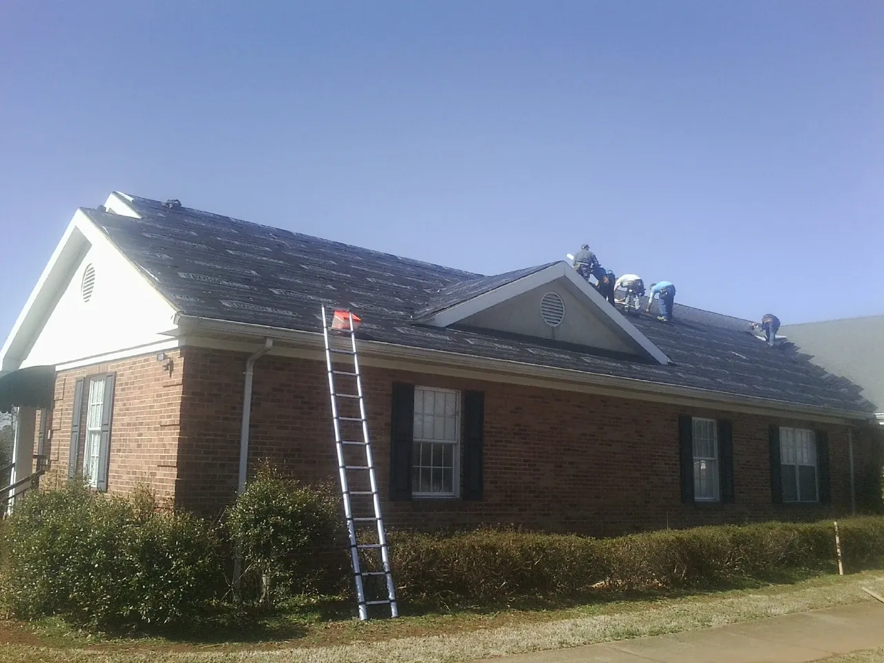 Crew tearing shingles off a brick ranch roof with a ladder set — Metal Roof Repair work by our crew in Arley