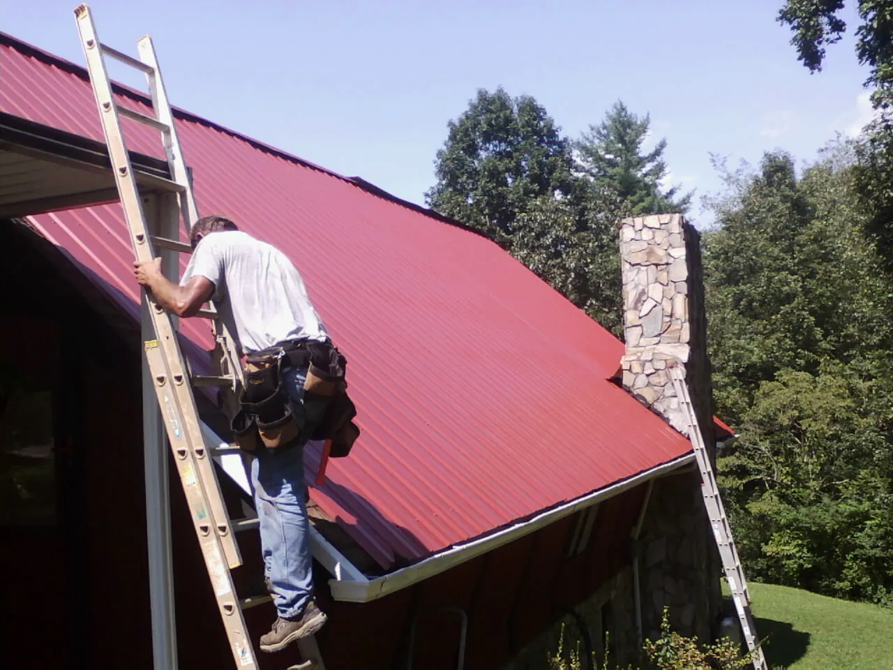 Roofer on a ladder beside a red metal roof and stone chimney — Soffit & Fascia Repair by Roofing Brothers in Arley